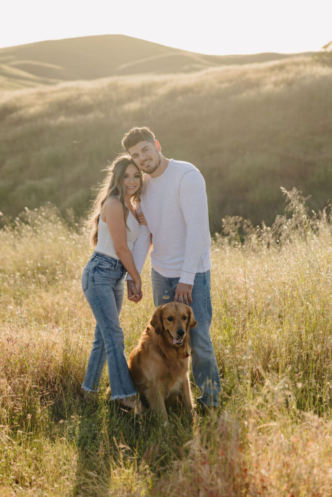 A couple holding hands and smiling with their dog sitting at their feet. 