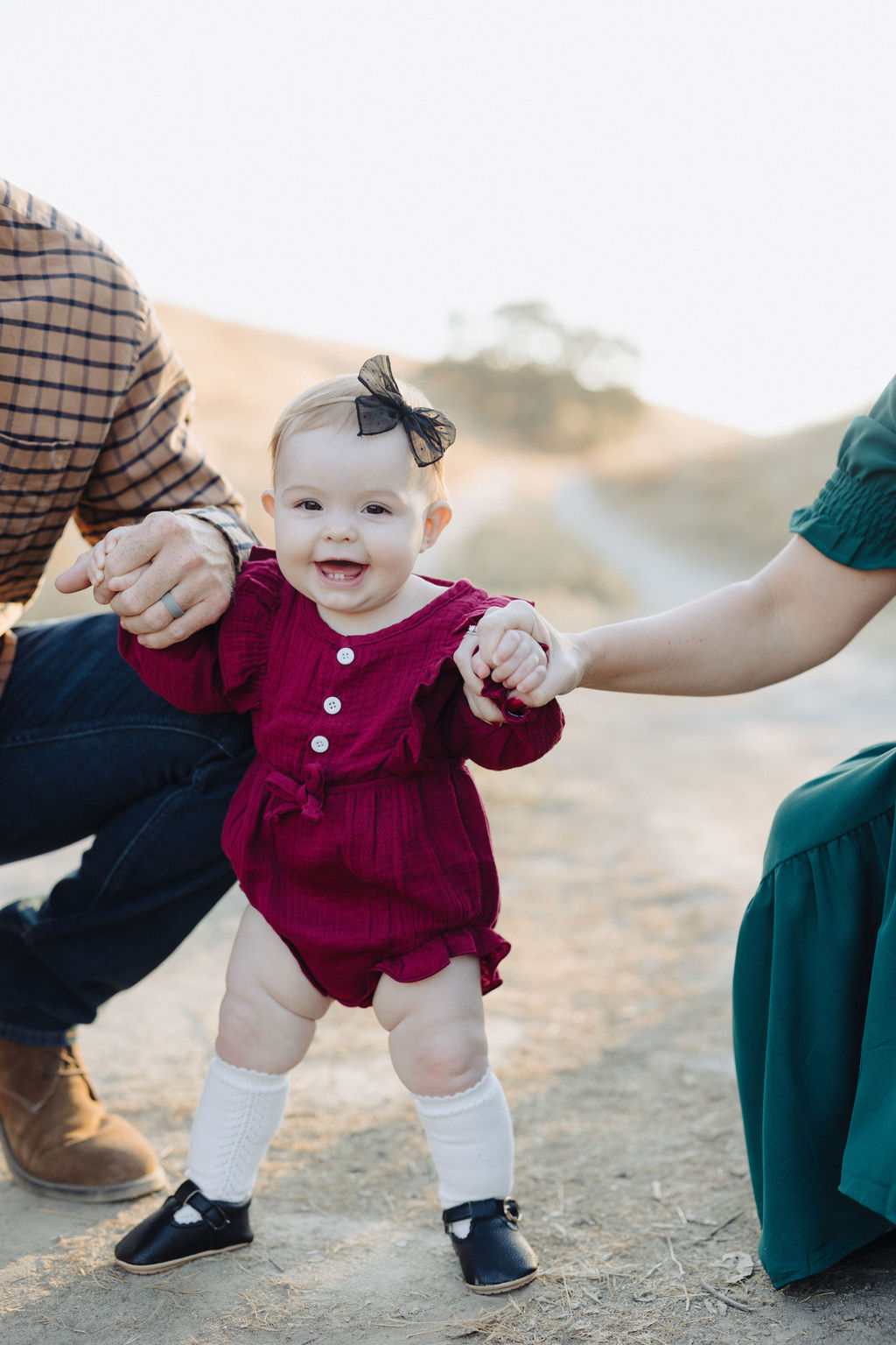 A baby standing on a dirt path holding both parent's hands